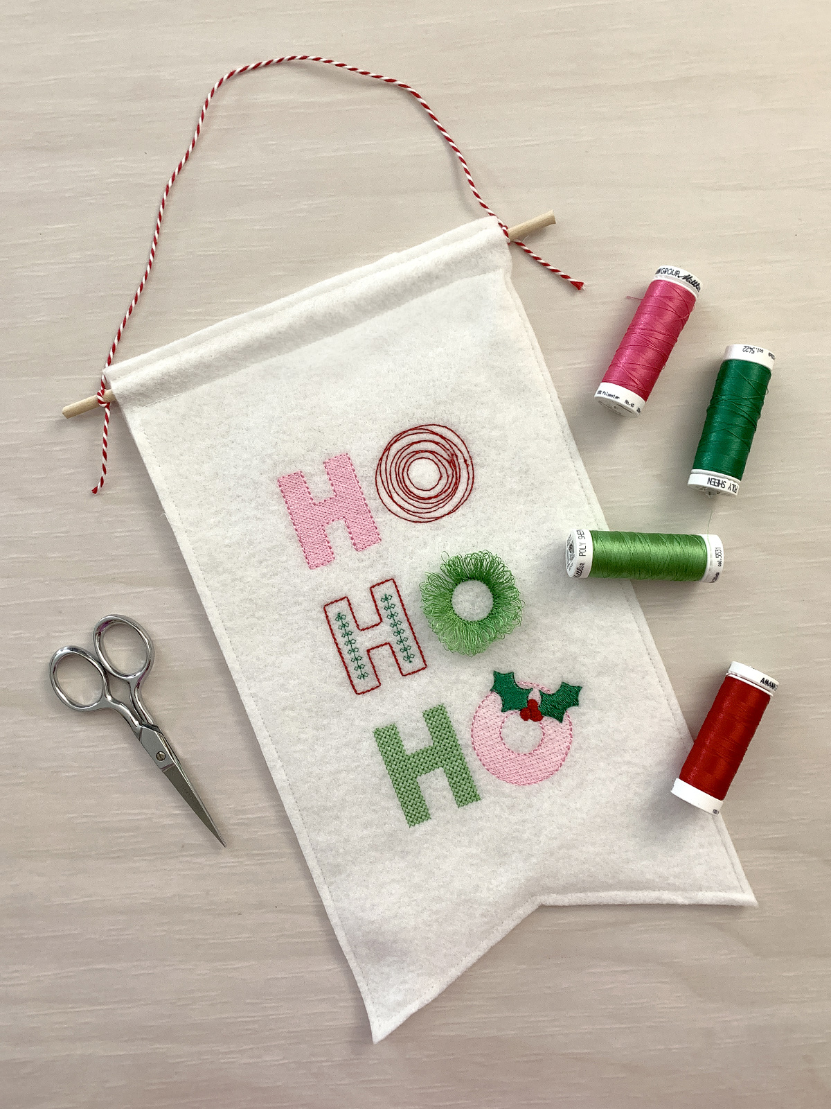 Small felt banner featuring embroidered letters spelling, "Ho! Ho! Ho!" The middle "O" is a special "fringe" stitch that adds a 3D effect that resembles a wreath when the bobbin threads are cut from the back side.