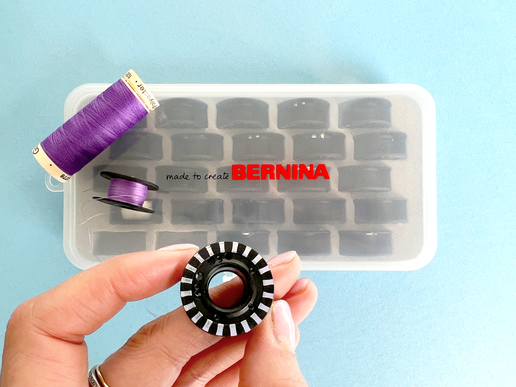 A had holds up a jumbo size black plastic bobbin. In the background is a case full of bobbins and a spool of sewing thread.