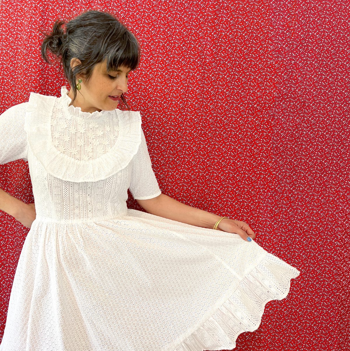 A dark haired woman is wearing a white eyelet dress with ruffles, against a red backdrop printed with tiny white flowers.