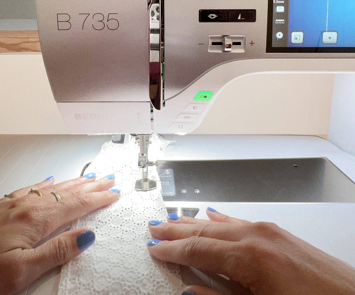 Two hands guide white fabric into a sewing machine that is fitted with a Gathering Foot.