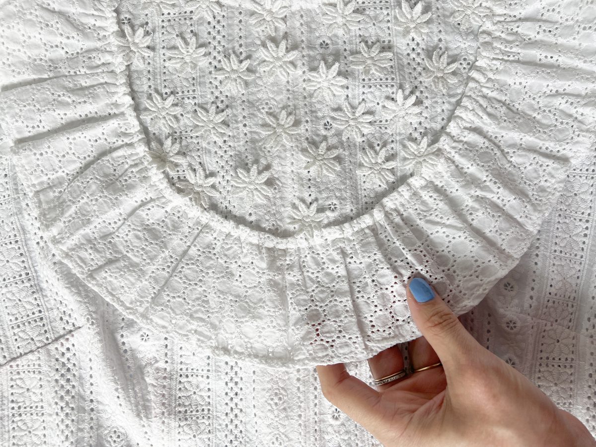 A hand is holding a curved ruffle attached to a white cotton dress. There are small flowers appliquéd above the ruffle.
