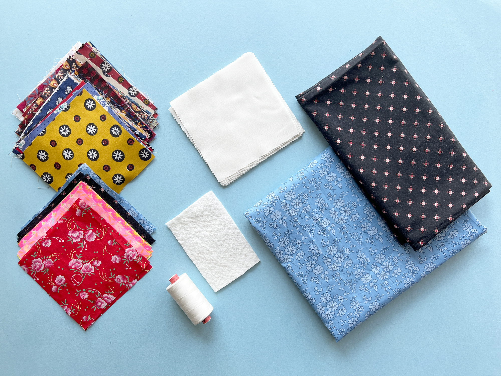 Squares of printed fabric and solid cream fabric, along with larger pieces of printed blue and black quilting cotton, along with a spool of thread are displayed against a light blue background.