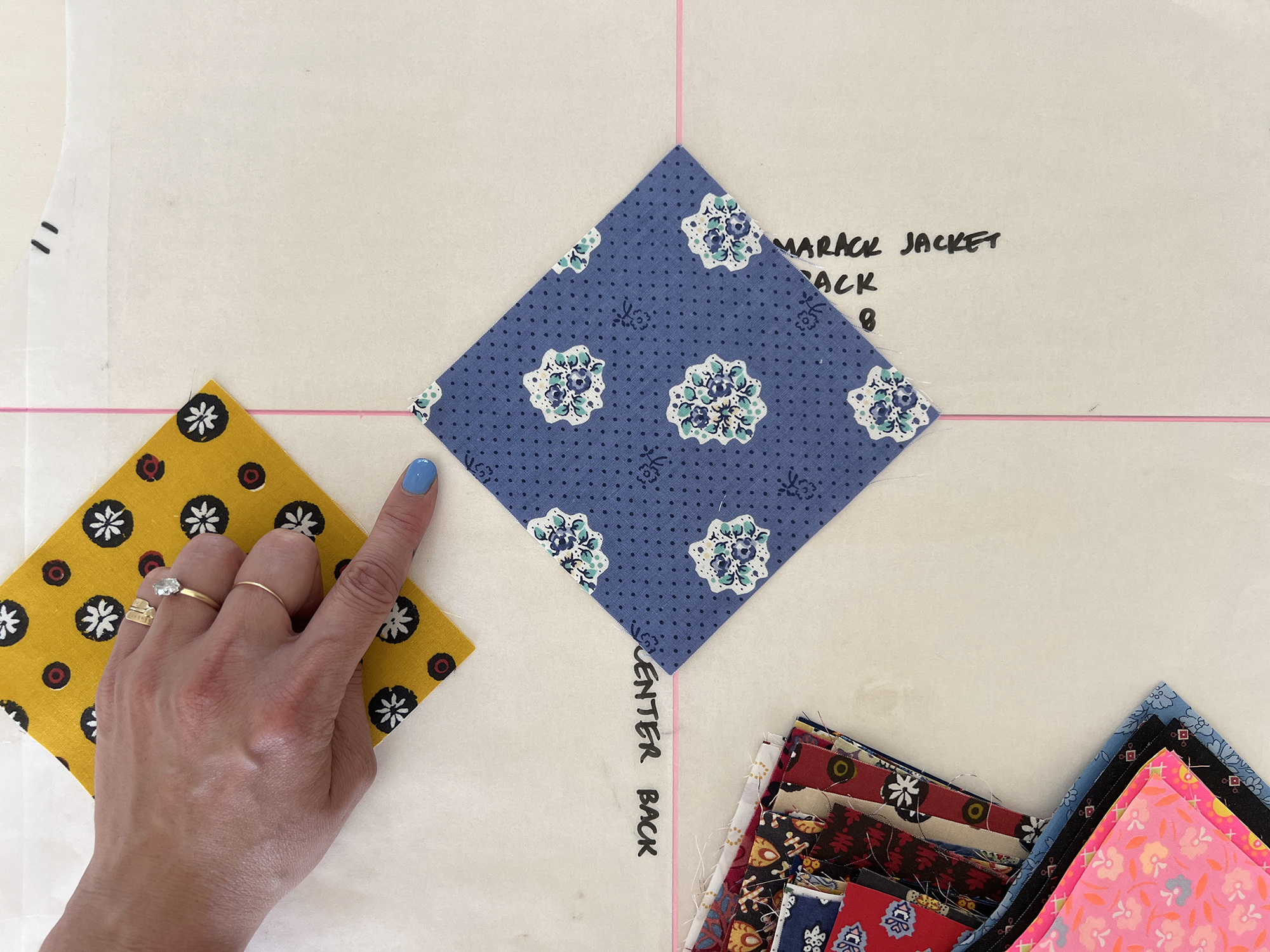 A blue square of fabric is placed so the corners match up with guide lines. A hand is pointing to the corner of the fabric.