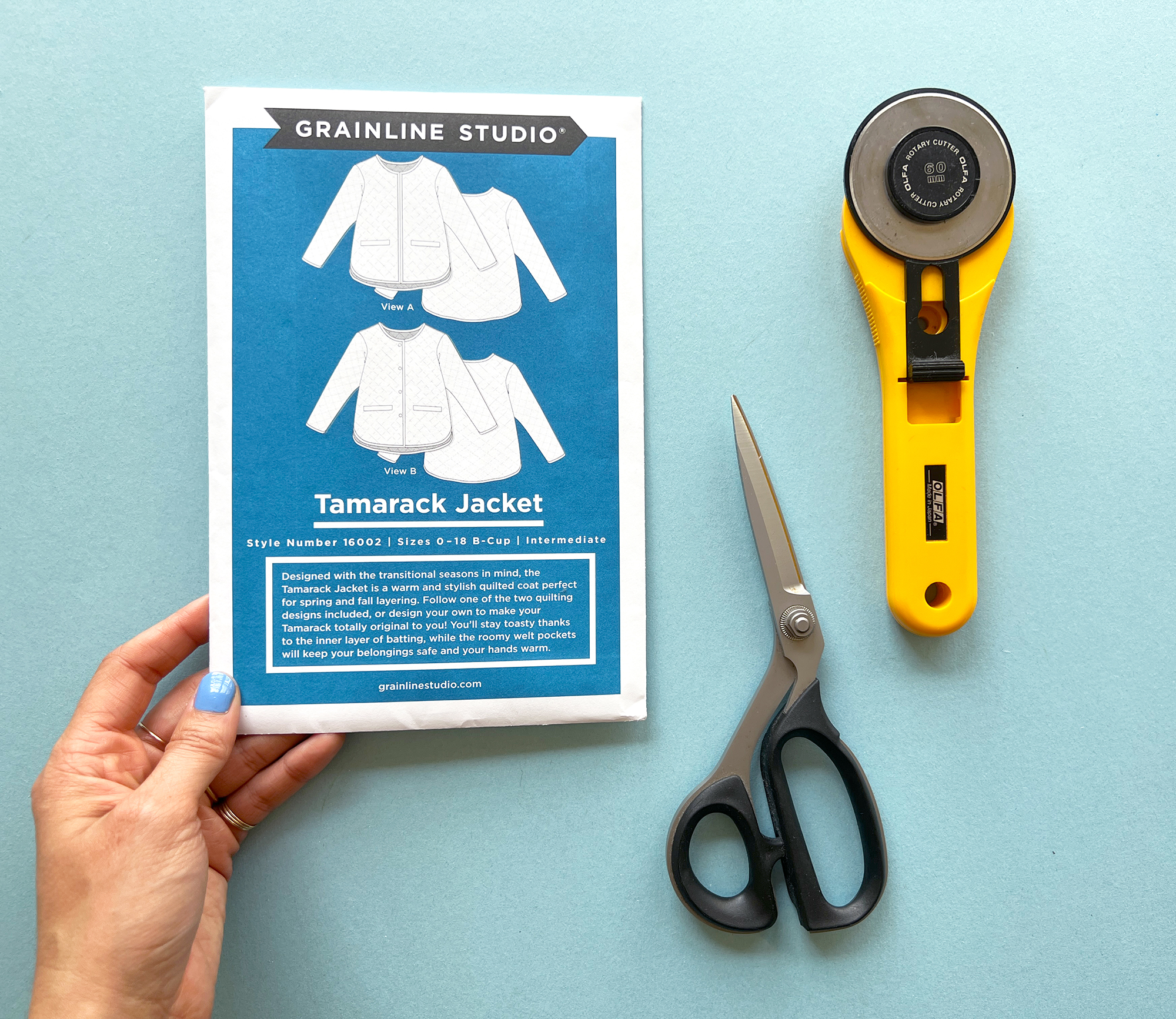 A hand is holding the sewing pattern envelope for Grainline Studio's Tamarack Jacket. There is also a rotary cutter and pair of scissors, all displayed on a light blue background.