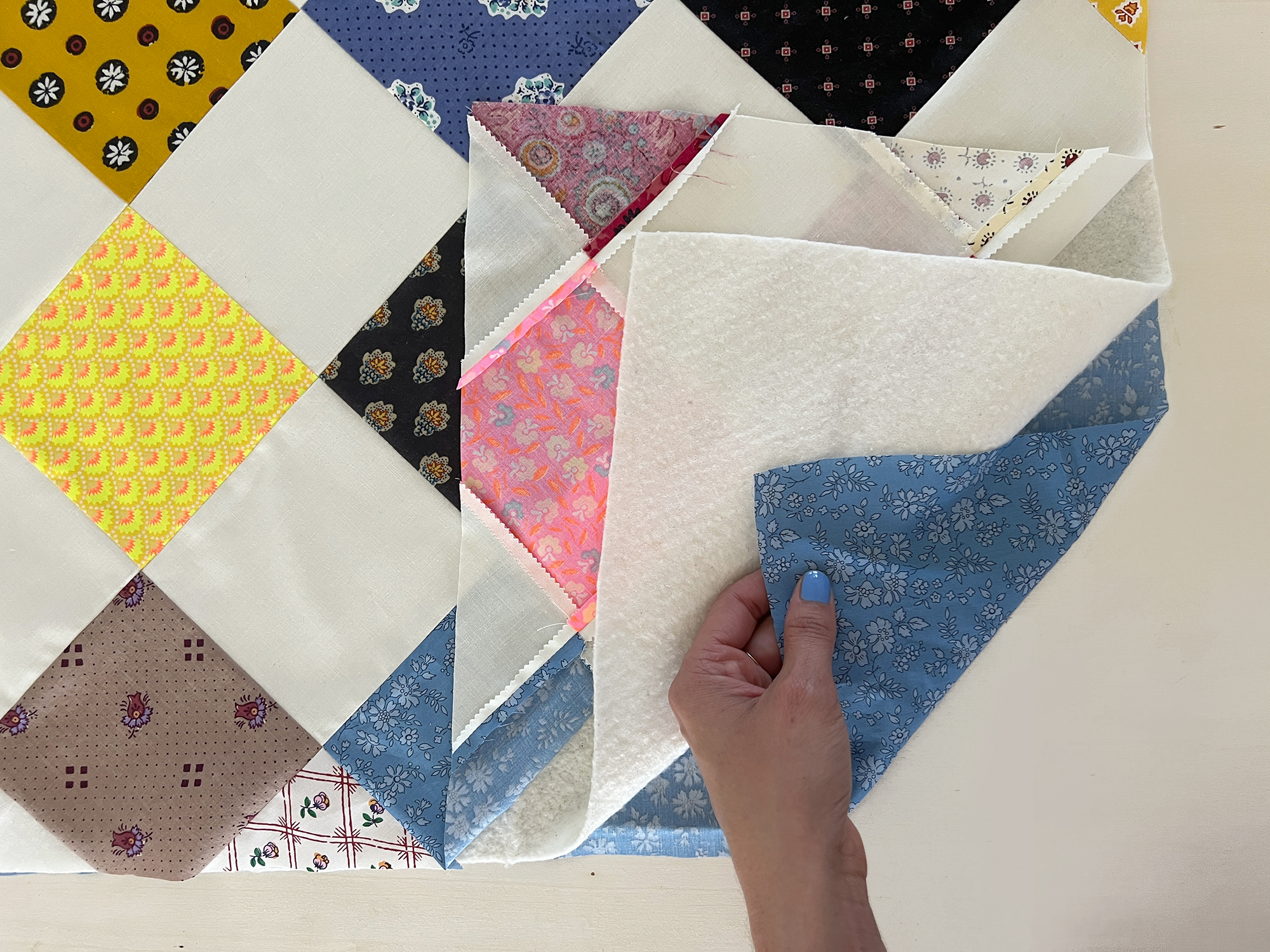 A hand flips through the layers of a quilt sandwich: lining on the bottom, cotton batting, and then checkerboard patchwork on the top layer.