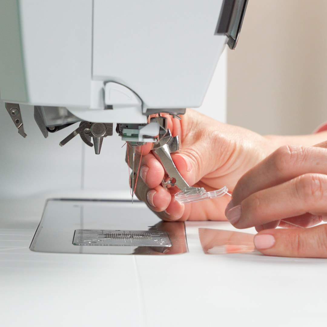 closeup view of a woman’s hands holding the Clear Embroidery Foot #39C under a sewing machine and inserting a white thread through it