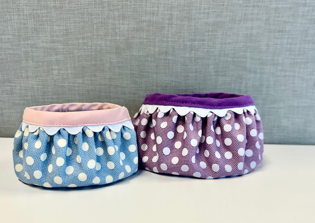 two finished fabric bowls in front of a gray background. One is blue and white polka dot with pink trim; the other purple polka dot with purple trim