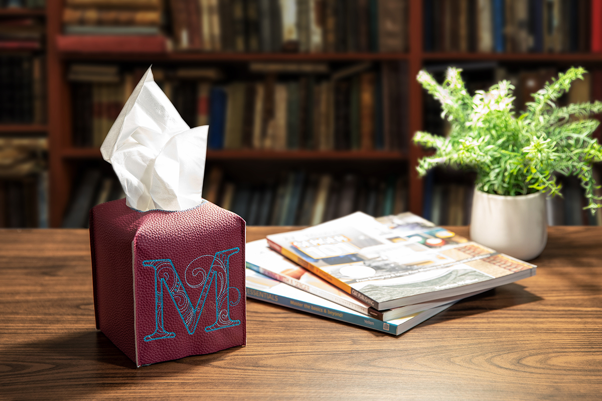 Brown sensory tissue box embroidered "M" on wooden desk with other items
