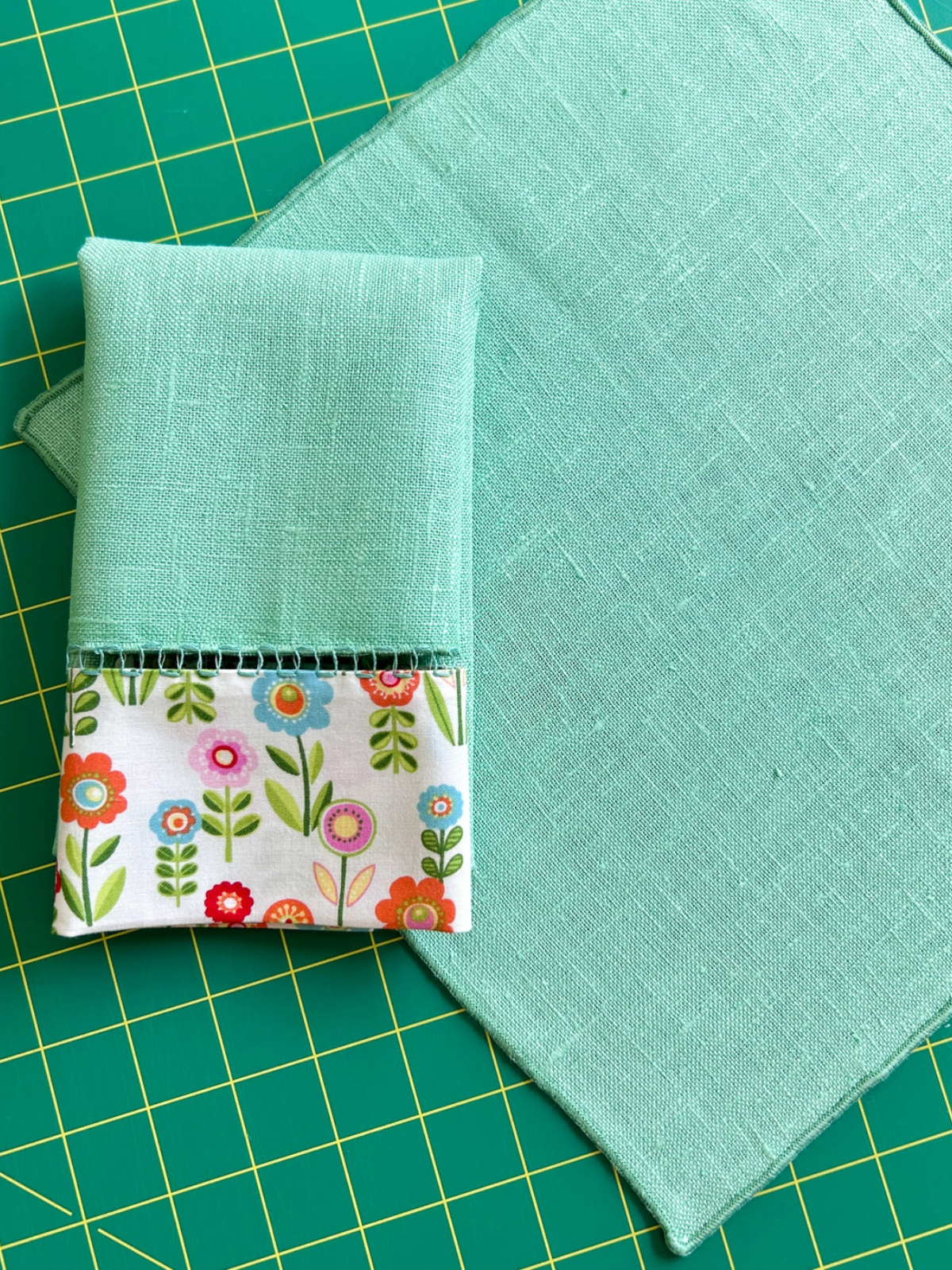 A folded green napkin laying on top of a plain green napkin square.