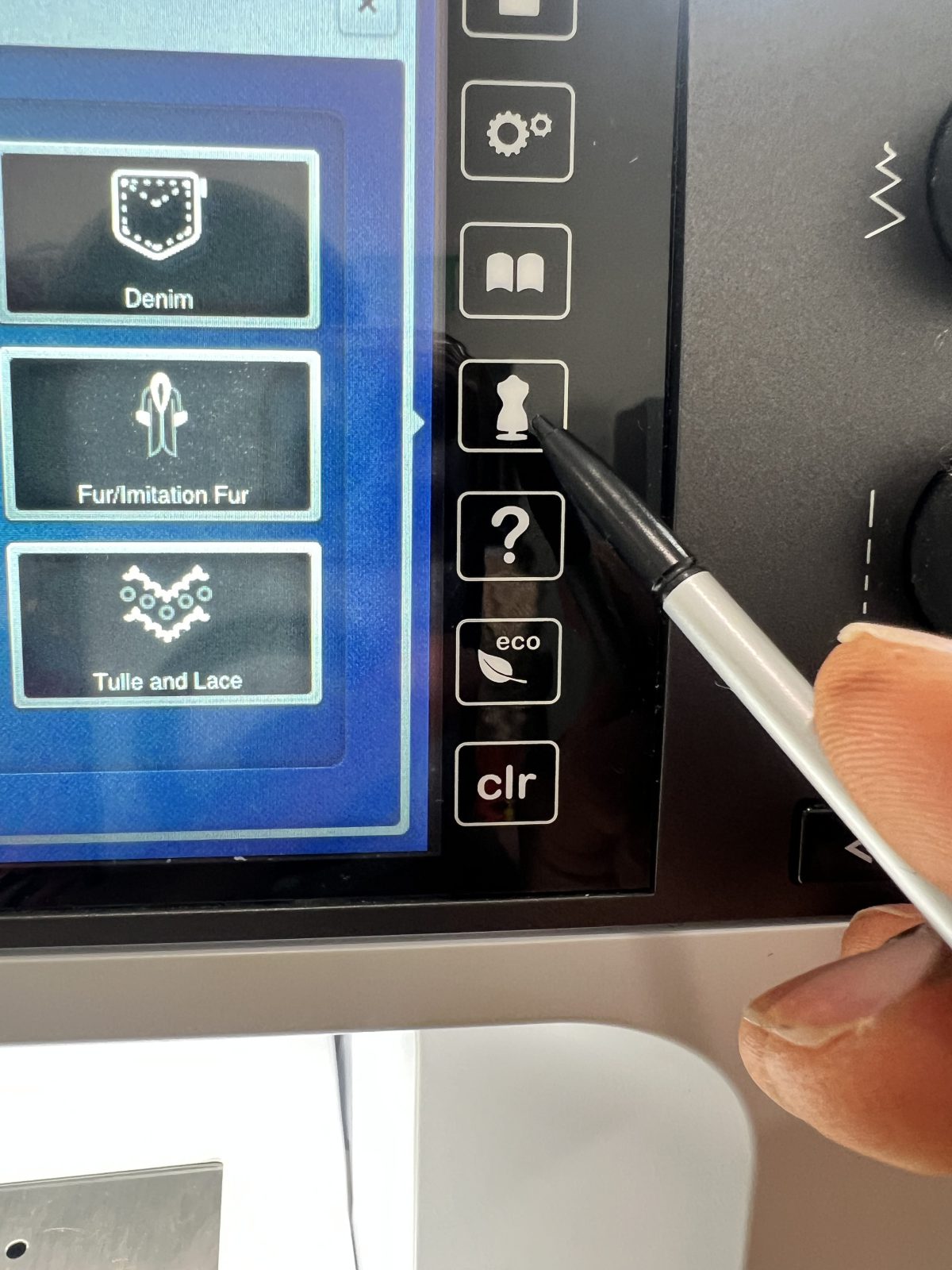 Close-up of a Bernina B 790 Pro sewing machine touchscreen. A hand holding a stylus is pressing the dress form icon on the right-side menu, with fabric options such as denim, fur/imitation fur, and tulle and lace visible on the screen