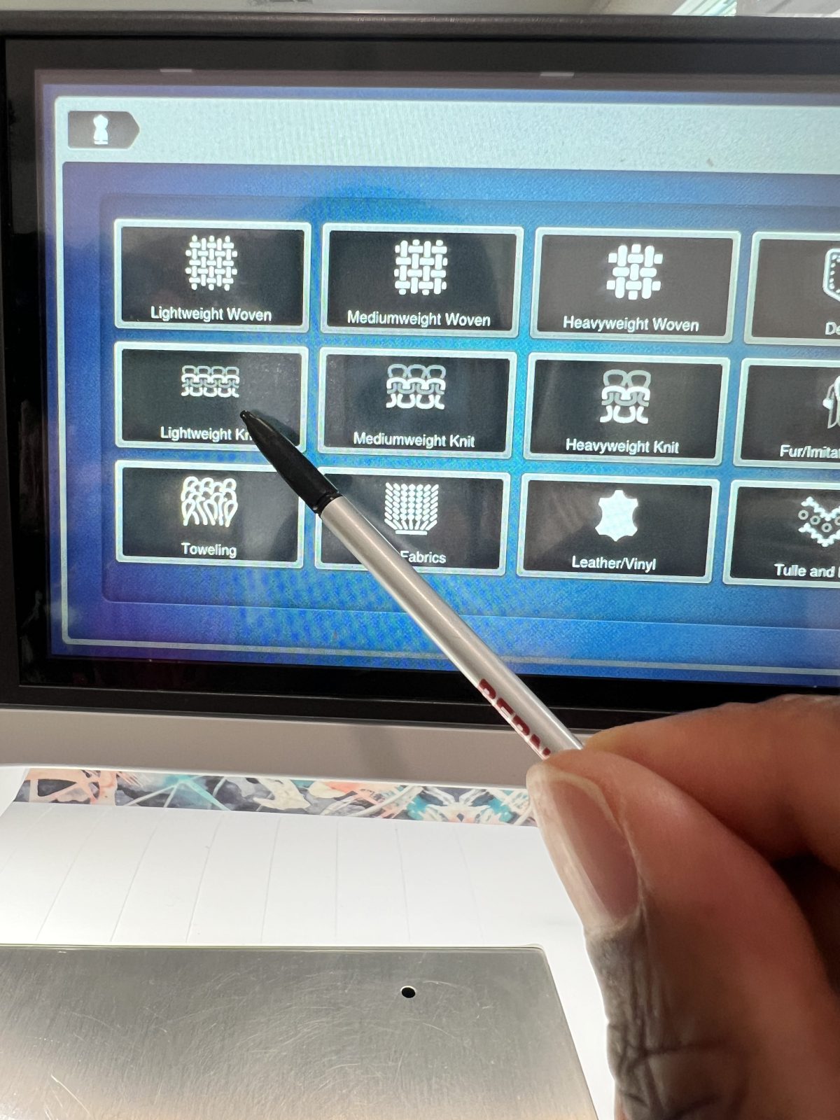 Close-up of a Bernina B 790 Pro sewing machine touchscreen. A hand holding a stylus is pointing to the ‘Lightweight Knit’ option on the fabric selection menu, which also displays choices for woven fabrics, mediumweight knit, heavyweight knit, toweling, leather/vinyl, and other materials.