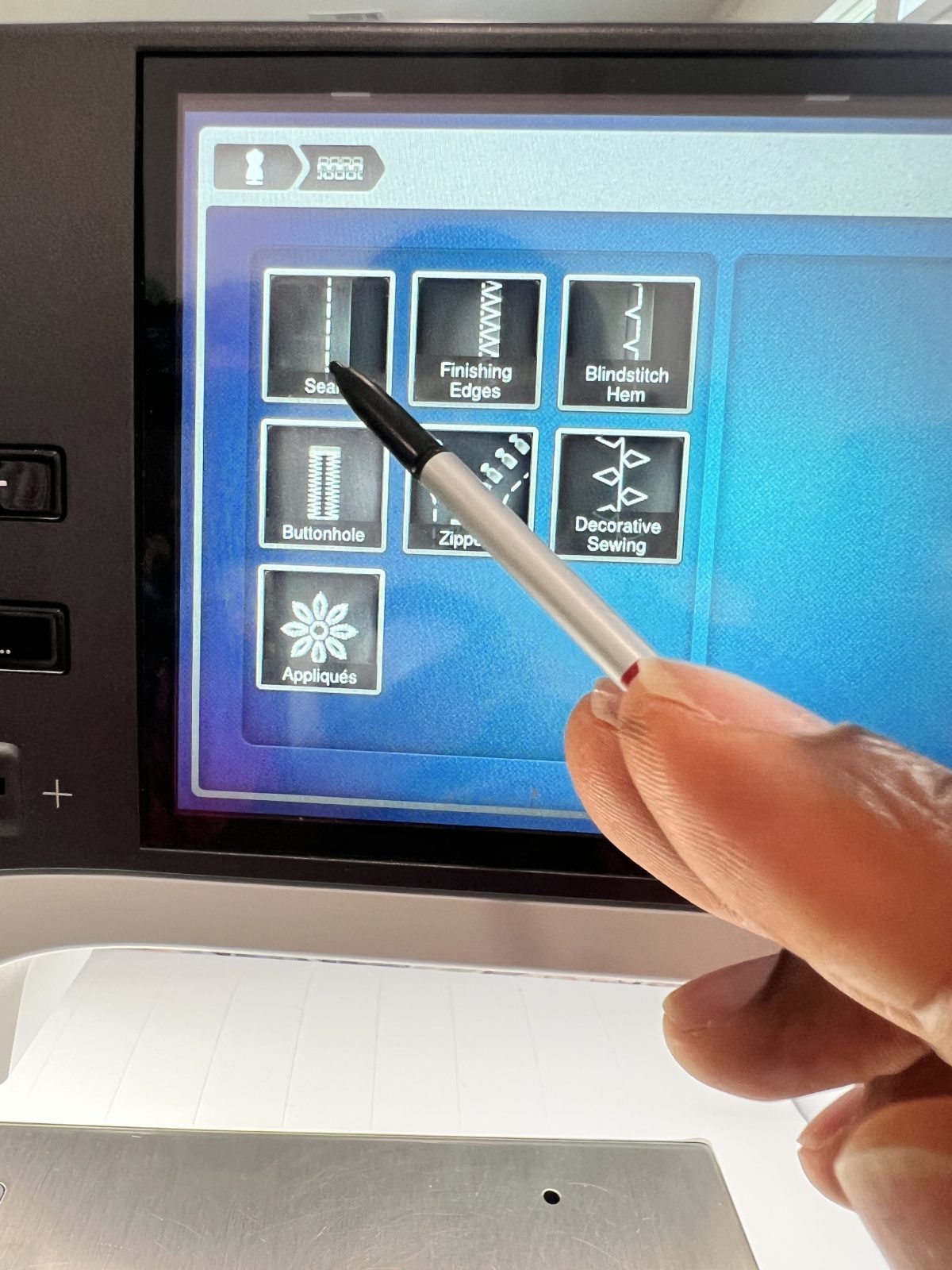 Close-up of a Bernina B 790 Pro sewing machine touchscreen. A hand is holding a stylus and pointing to the ‘Seam’ option on the menu, which also shows icons for finishing edges, blindstitch hem, buttonhole, zippers, decorative sewing, and appliqués