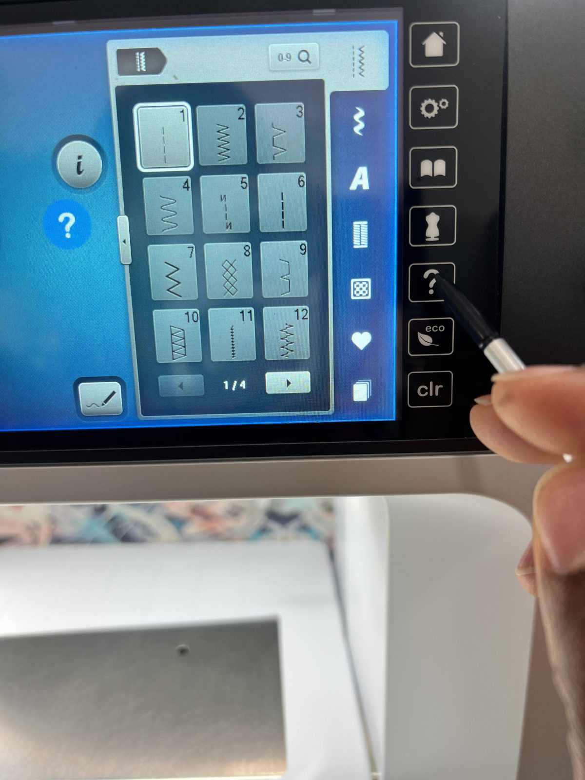 Close-up of a sewing machine touchscreen showing a menu of stitch options, including straight and zigzag stitches. A hand is using a stylus to press the question mark button on the side panel to access stitch information.