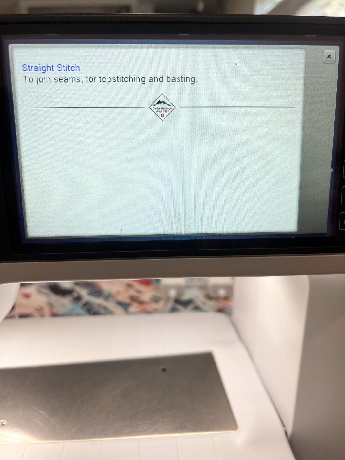 Close-up of a sewing machine screen displaying stitch information. The screen shows ‘Straight Stitch: To join seams, for topstitching and basting’ with a small Swiss Heritage logo beneath the text. The metal needle plate and part of the sewing surface are visible below the screen.