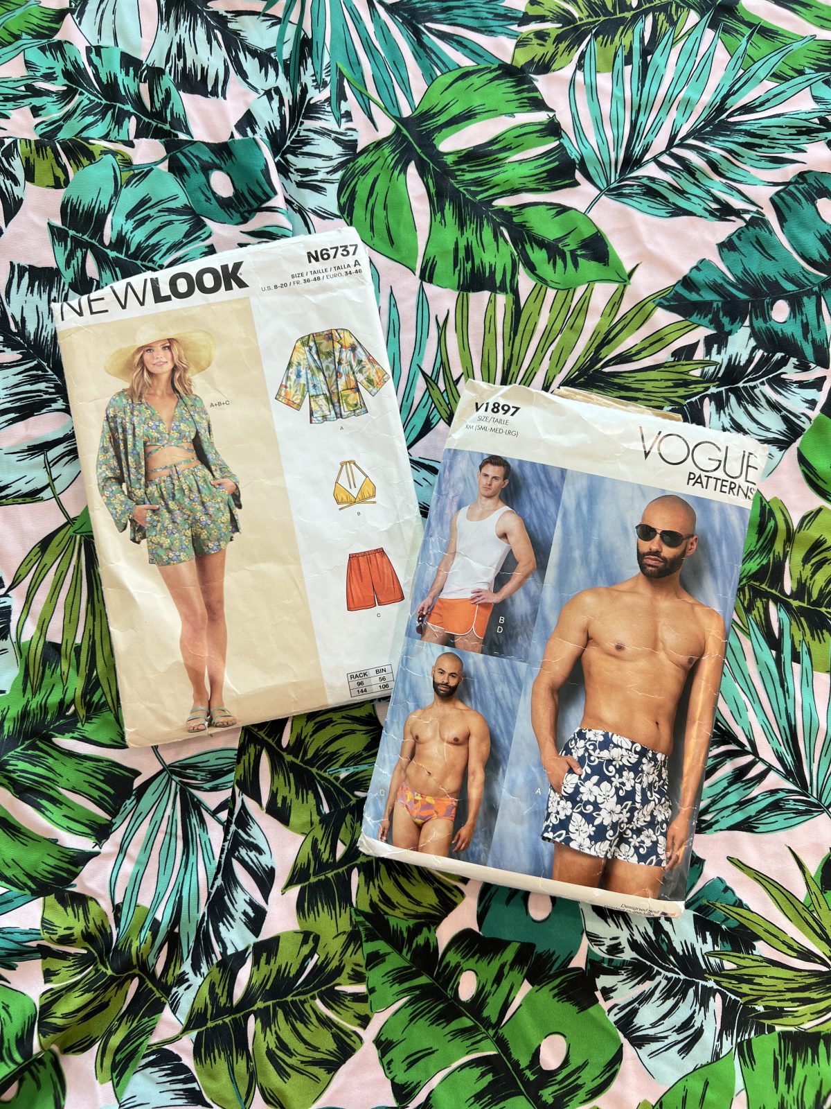 Two sewing pattern envelopes, New Look 6737 and Vogue 1897, placed on top of tropical leaf print fabric in green, teal, black, and pink. The New Look pattern shows a woman in a matching jacket, bikini top, and shorts, while the Vogue pattern shows men modeling various swim trunks and shorts.