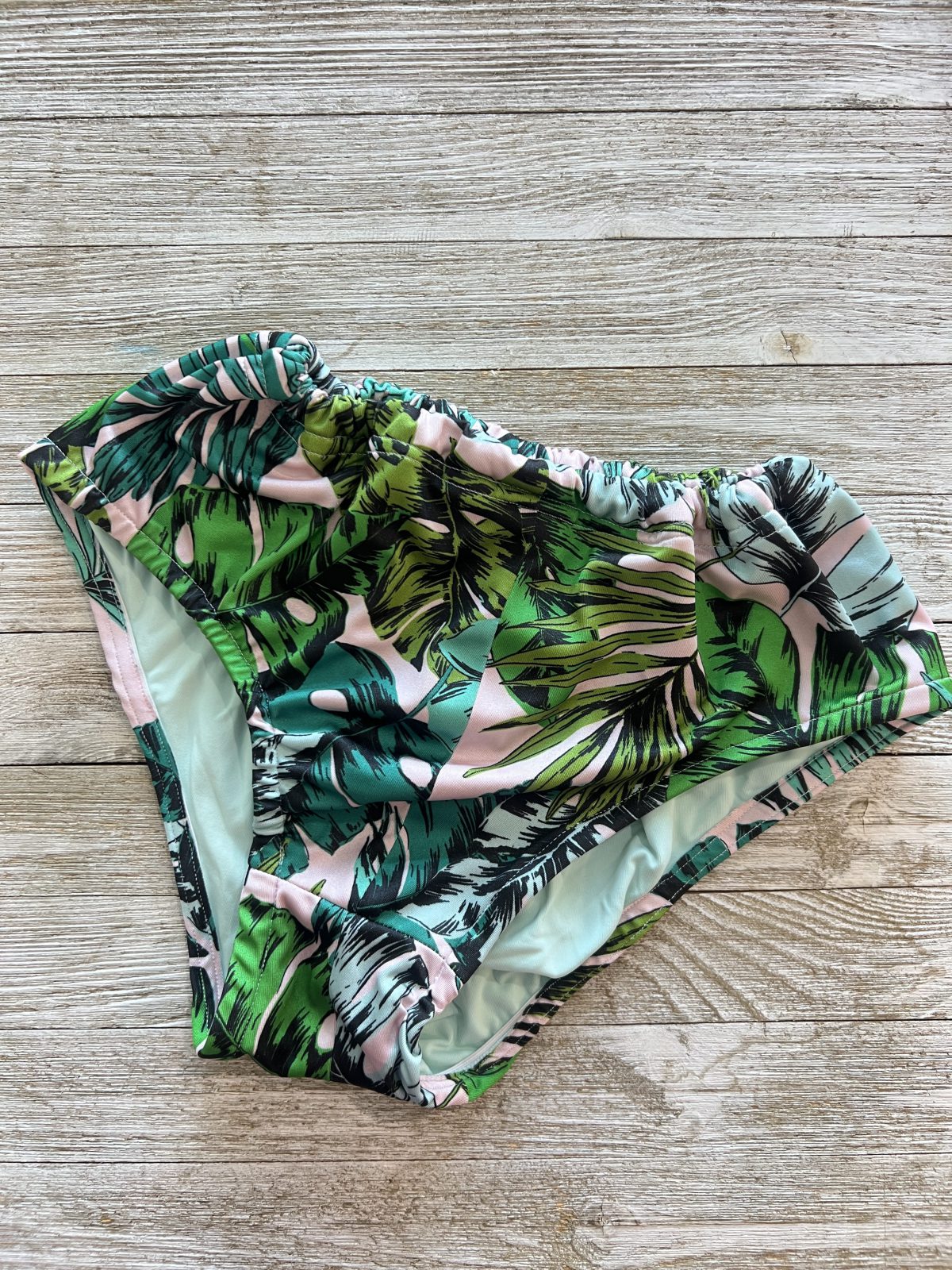 High-waisted bikini bottoms with a tropical leaf print in shades of green, teal, black, and pink, displayed on a wooden surface