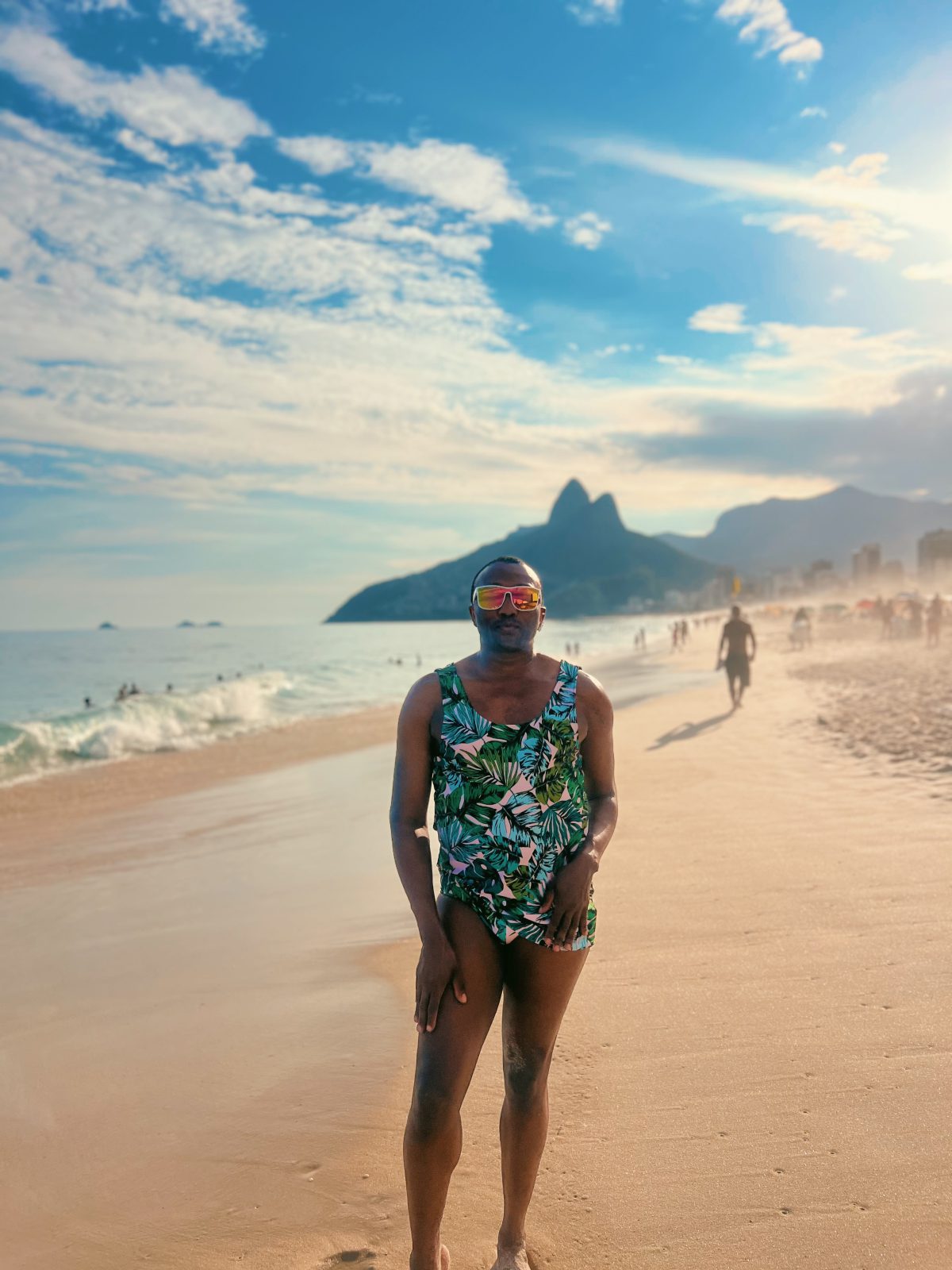 Person standing barefoot on a sandy beach at sunset, wearing a tropical green, teal, black, and white leaf-print swimsuit with matching top and bottoms. They are wearing reflective sunglasses and posing with one hand resting by their side. Ocean waves, beachgoers, and distant mountains are visible in the background under a partly cloudy sky.