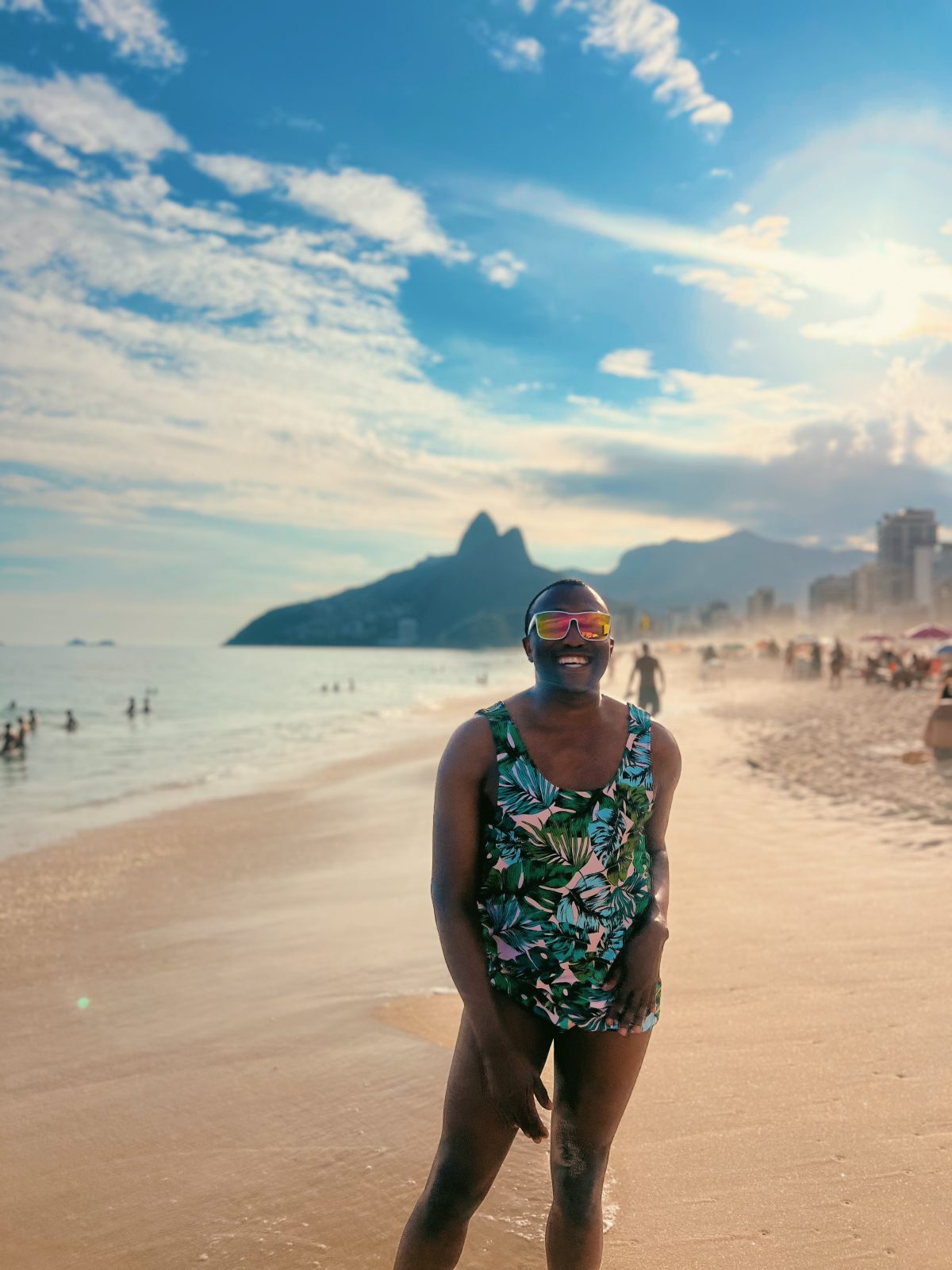Person standing barefoot on a sandy beach at sunset, wearing a tropical green, teal, black, and white leaf-print swimsuit with matching top and bottoms. They are wearing reflective sunglasses and posing with one hand resting by their side. Ocean waves, beachgoers, and distant mountains are visible in the background under a partly cloudy sky.