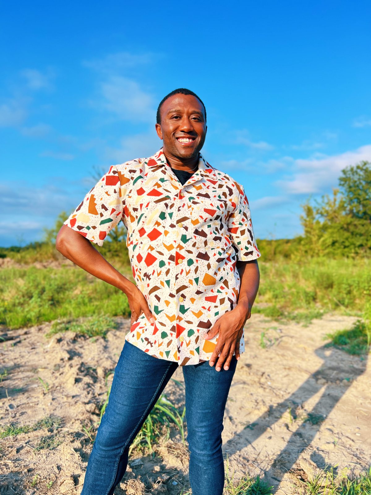 Person standing outdoors in a colorful handmade button up shirt with a terrazzo inspired print, paired with blue jeans, smiling under a bright blue sky