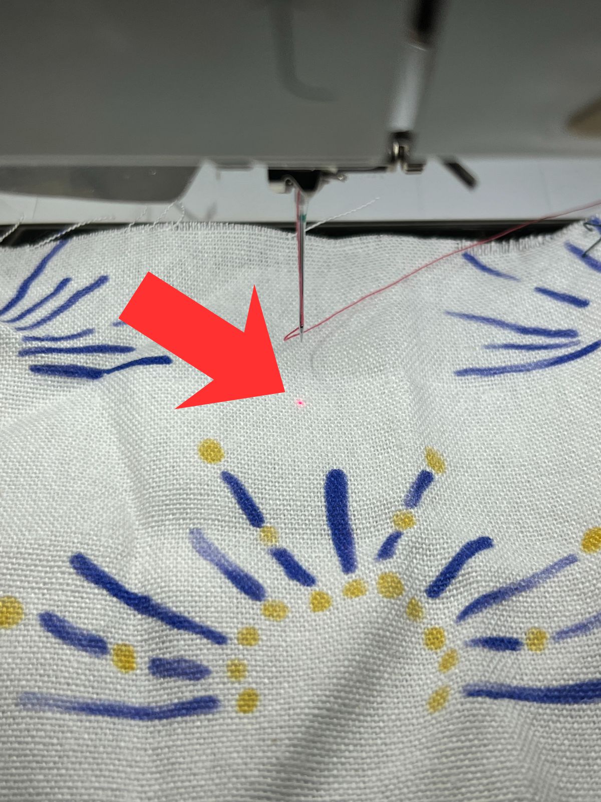 Close-up of fabric under a sewing machine needle with a small red laser point marking the stitch path. The fabric is white with a blue and yellow abstract print, and a red arrow is pointing toward the laser dot.