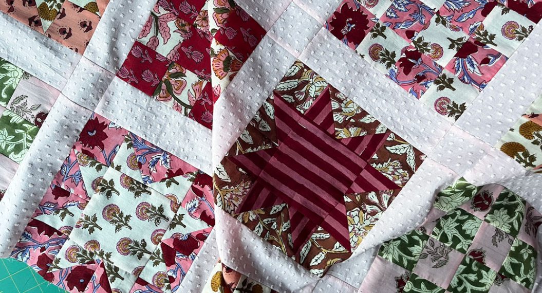 A close up of a quilt in progress. The quilt features block prints arranged in alternating quilt blocks of 16-patches and sawtooth stars. The colors are mostly in shades of pink and red.