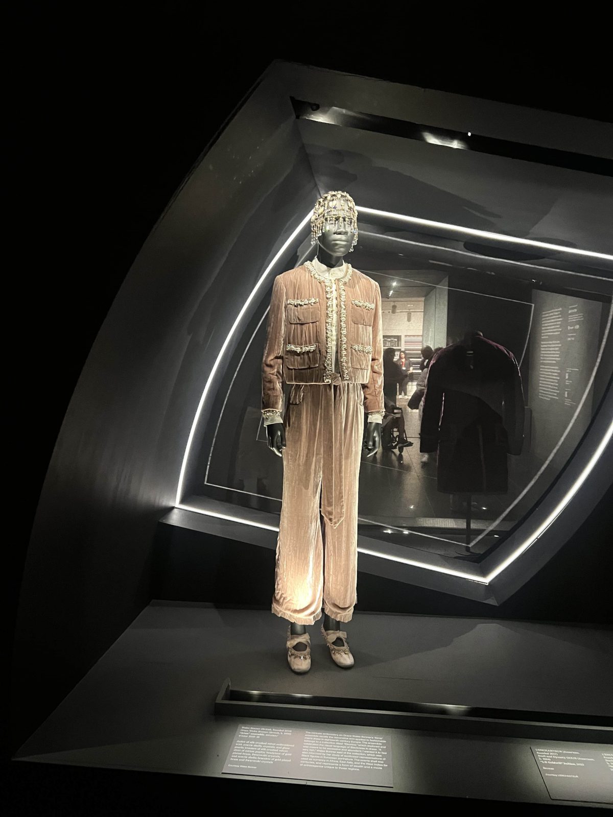 A mannequin in a museum display case wearing a soft beige velvet ensemble with a cropped jacket, matching wide pants, and a beaded headpiece with strands falling around the face. The outfit is lit dramatically within a dark room.