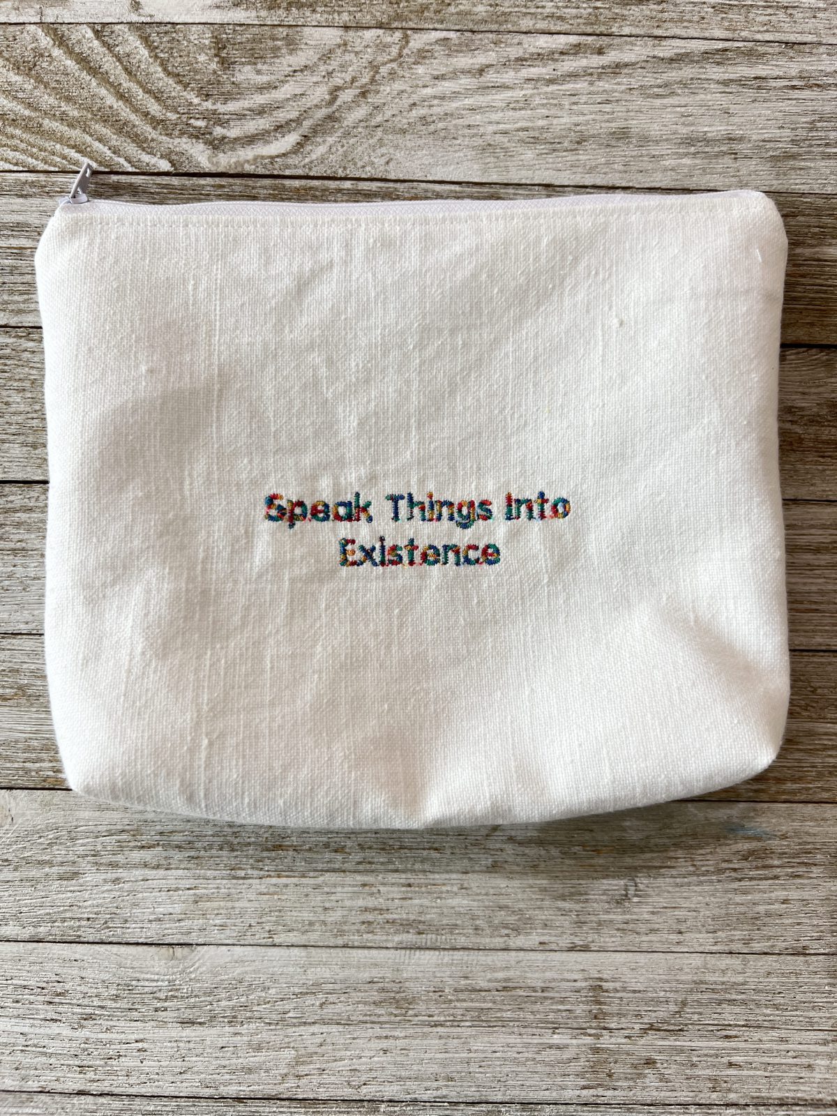 A white fabric zippered pouch lying on a wooden surface, with the phrase “Speak Things Into Existence” embroidered in multicolored thread in the center.