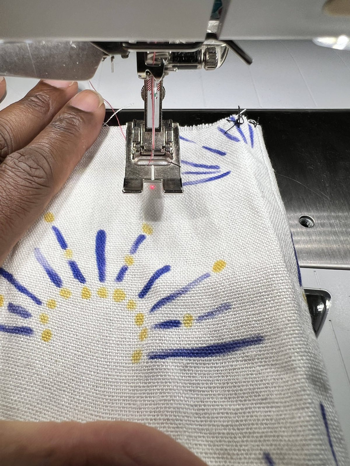 A close-up of a sewing machine needle and presser foot stitching white fabric with a blue and yellow abstract print. A small red laser dot marks the stitch line, and a hand is guiding the fabric.