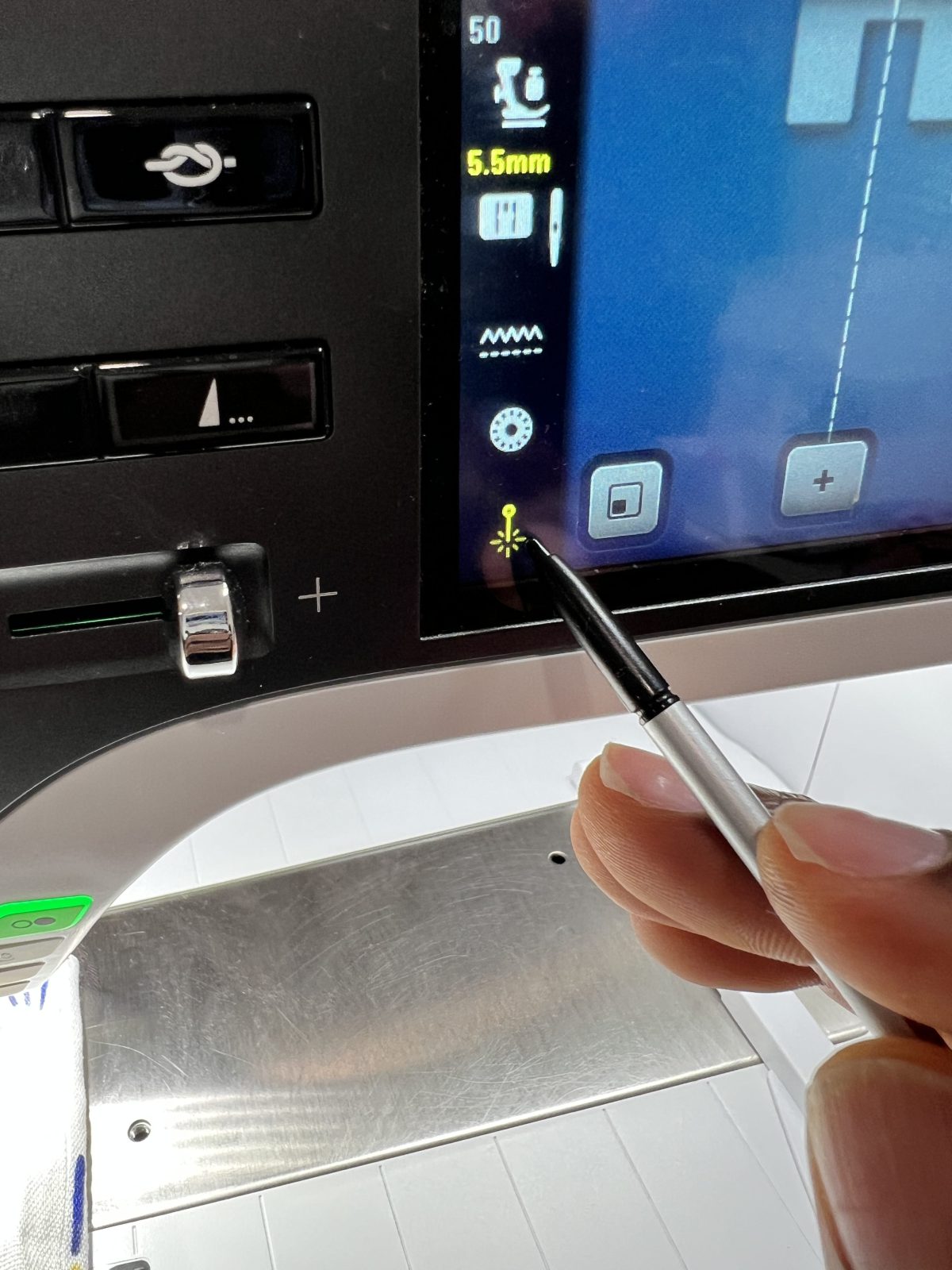 A close-up of a hand using a stylus to tap the laser guide icon on the touchscreen of a BERNINA B 790 PRO sewing machine. The machine screen displays stitch settings, and the workspace is visible beneath the needle.