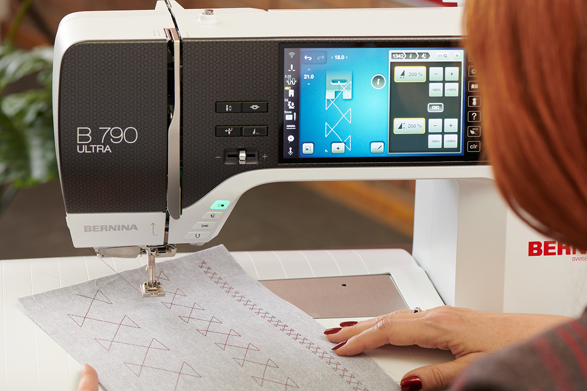 A sewing machine showing a large, geometric decorative stitch pattern on screen. Underneath the presser foot of the machine is a grey piece of fabric showing the pattern sewn out at 3 different sizes.